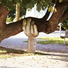 A tree with a sculpted stone hand supporting its bent trunk in a park setting.